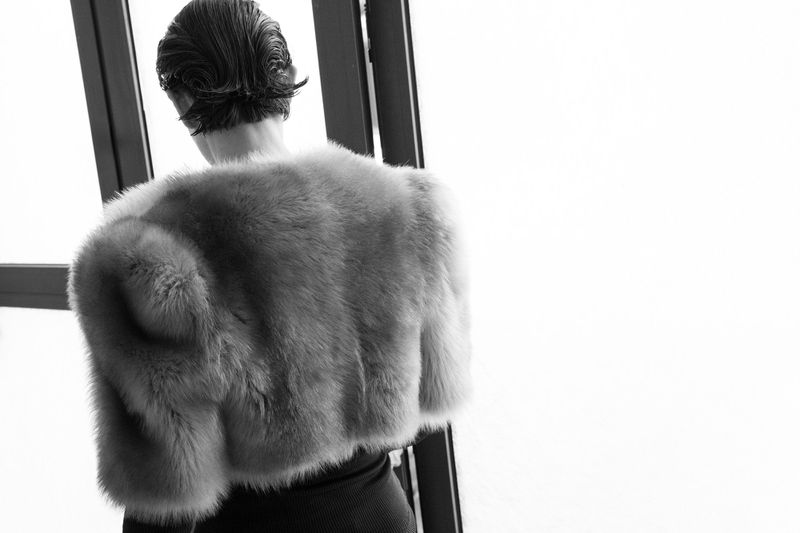 Black and white shot of an adult female with short, slicked-back hair wearing a fuzzy fur jacket with her back to the camera.