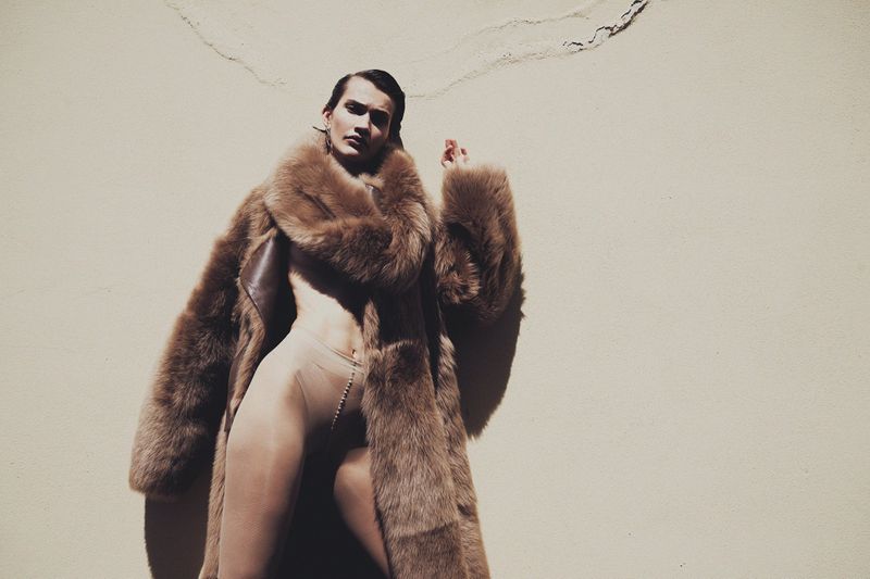 Adult in fur coat posing in front of wall in photo studio. Minimalist fashion photograph featuring earth tones and single human.