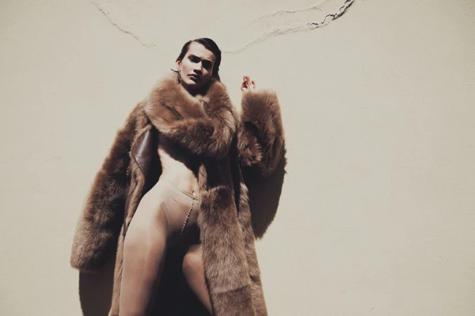 Adult in fur coat posing in front of wall in photo studio. Minimalist fashion photograph featuring earth tones and single human.