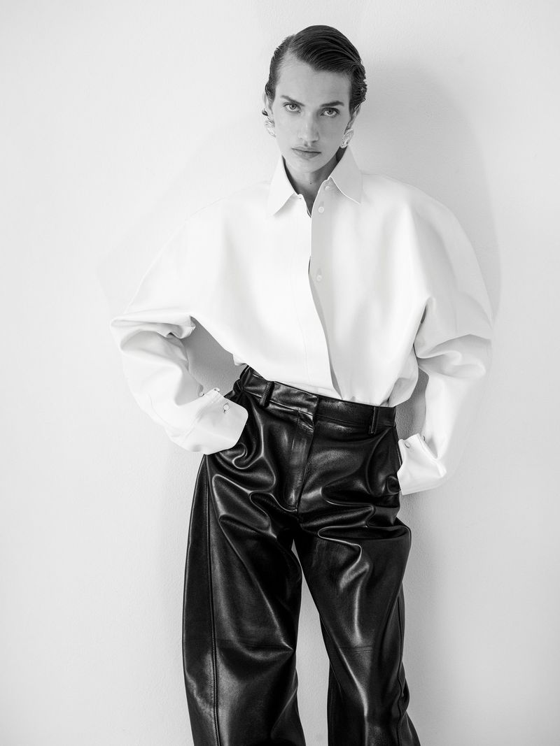Fashionable model poses in white shirt and black pants against a plain background. Studio lighting highlights contemporary clothing.