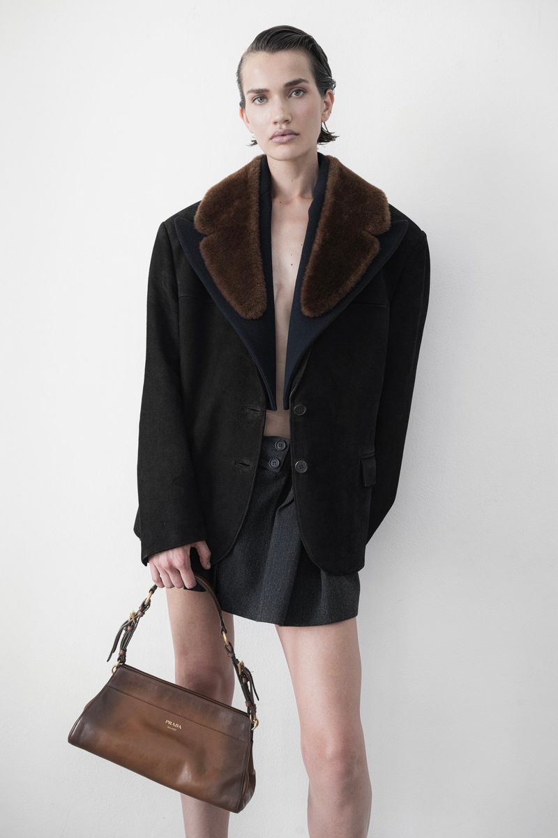 Model wearing a black jacket with a brown fur collar holding a brown leather handbag against a white studio backdrop.