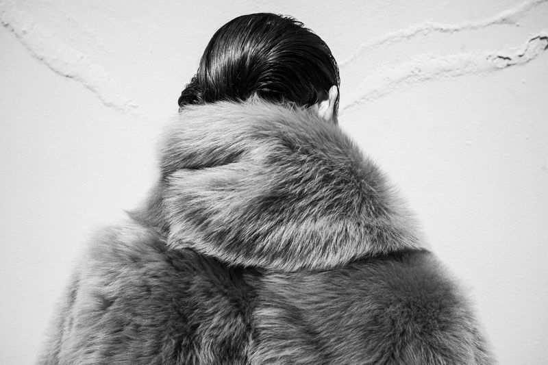 A woman in a large grey fur coat stands against a light grey textured wall, only the back of her head and coat are visible.