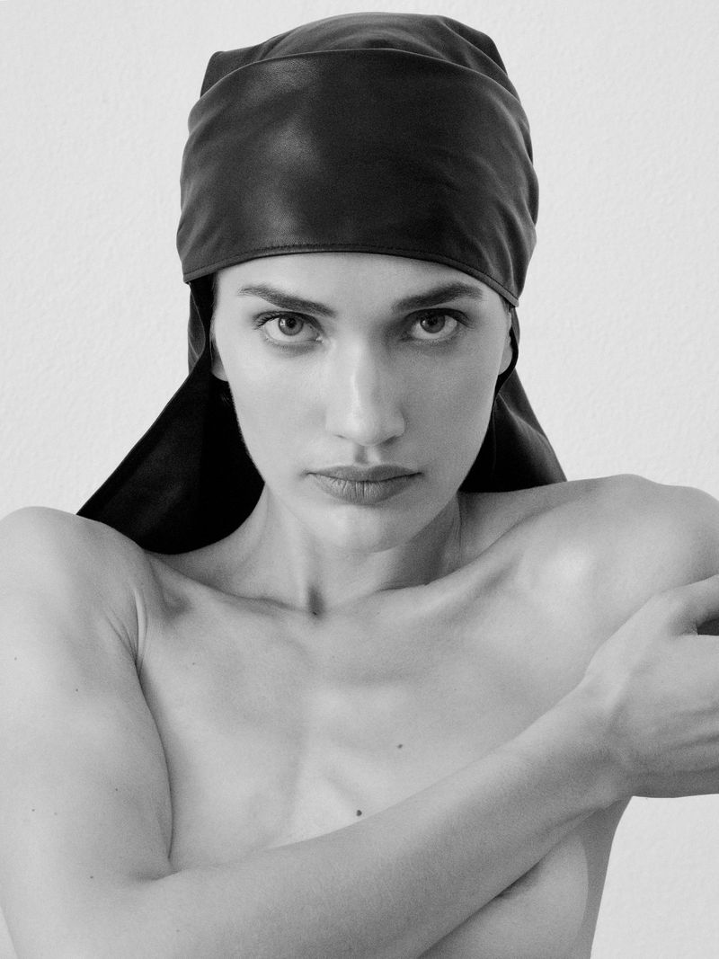 Black and white portrait of a woman with fair skin, wearing a black leather head covering and posing with arms crossed in front of a plain background.