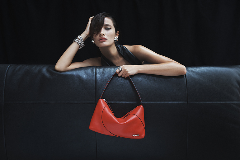 A fashionable adult female model wearing a black dress lies on a black couch with a red Mimco purse against a dark backdrop.