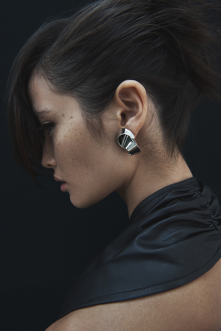 Close-up of a young adult woman in profile with dark hair, wearing large silver earrings and a black garment against a black backdrop.