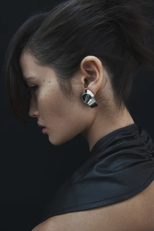 Close-up of a young adult woman in profile with dark hair, wearing large silver earrings and a black garment against a black backdrop.