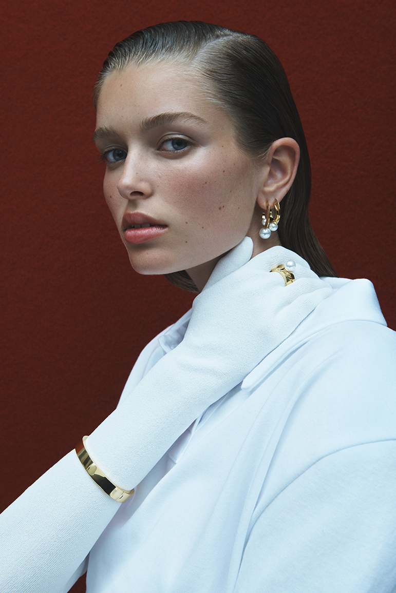 Close-up of a woman with slicked-back hair wearing gold jewelry, including earrings and a cuff bracelet, against a red background.