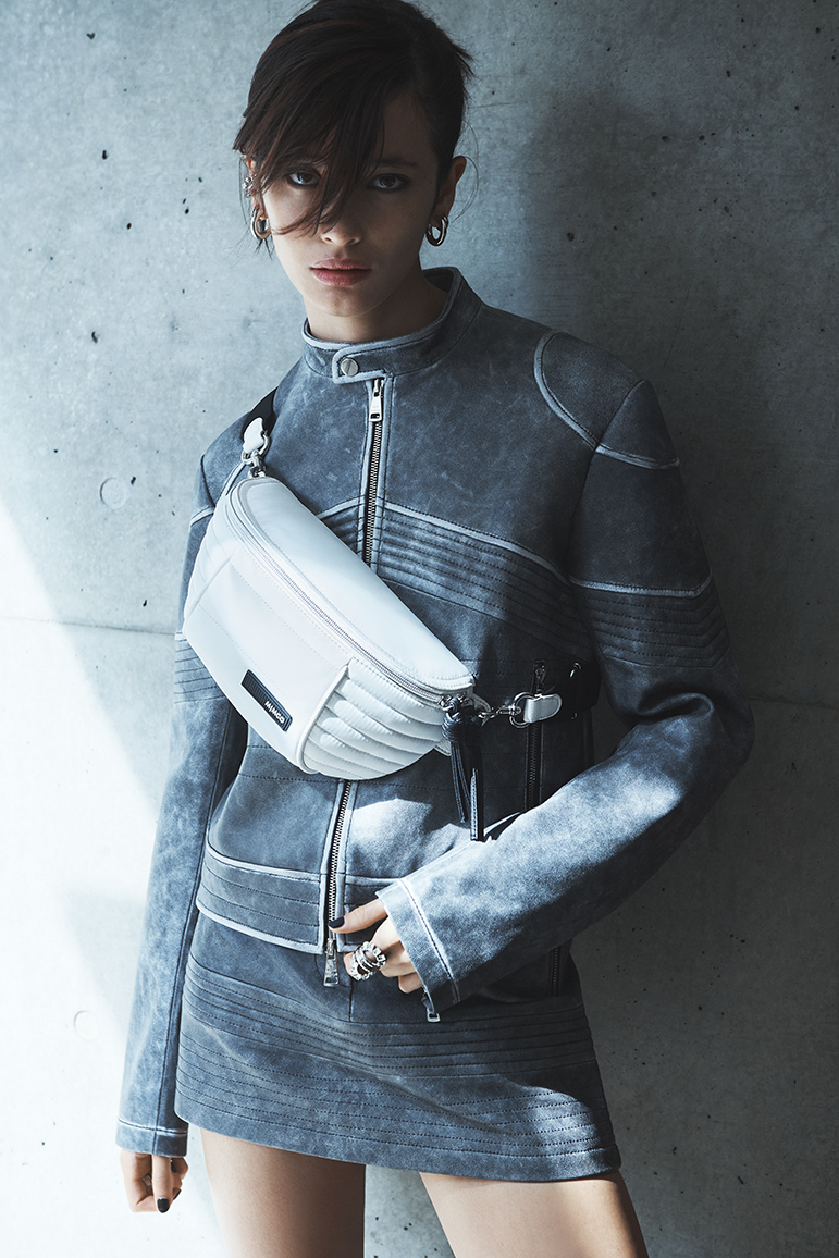 Portrait of a woman in a distressed denim jacket and skirt, wearing a white belt bag, posing against a concrete wall for a fashion shot.
