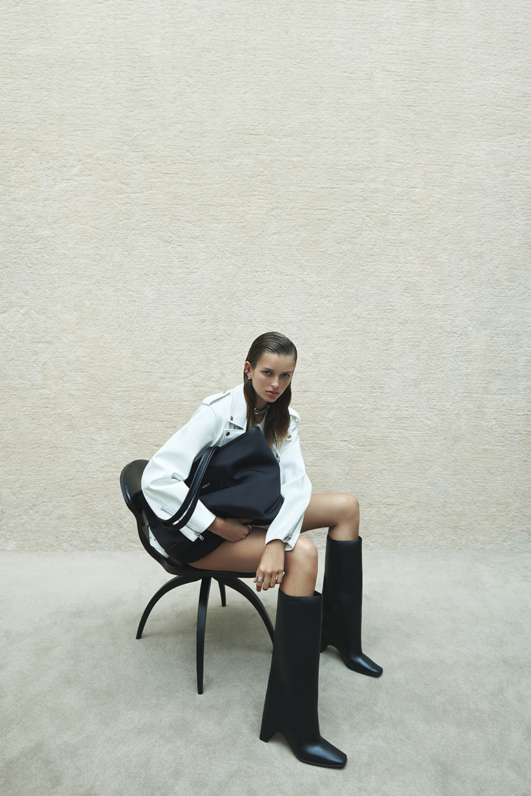 Woman in white jacket and black boots seated on a black chair holding a dark bag against a textured neutral wall