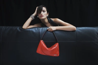 Woman in dark clothing holding a bright red handbag, leaning against a black leather sofa with silver bracelets.