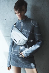 Model wearing a grey jacket and matching skirt holding a white crossbody bag against a textured concrete wall.