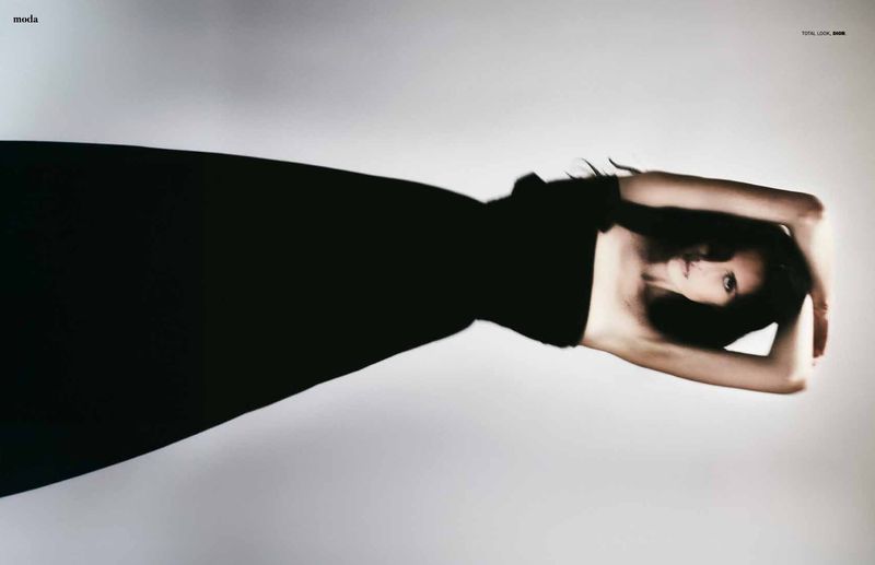 A fashion shot presents a woman with dark hair in a black dress against a soft grey backdrop, arms above her head in a striking pose.