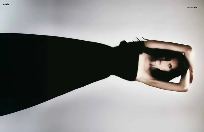 A fashion shot presents a woman with dark hair in a black dress against a soft grey backdrop, arms above her head in a striking pose.