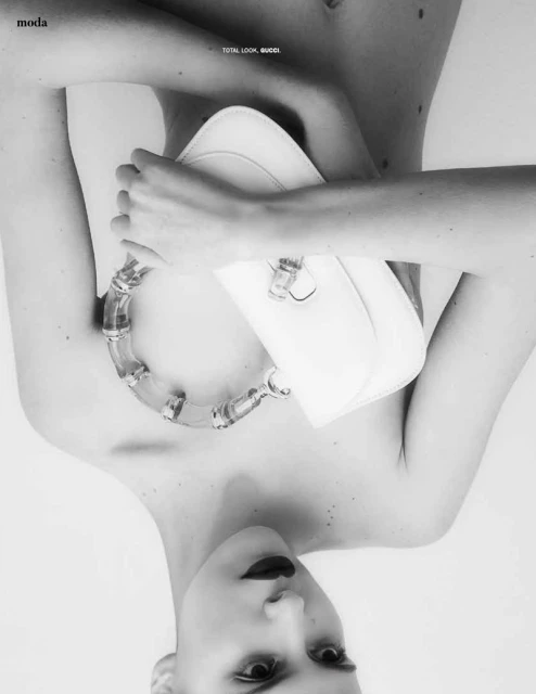 A person faces the camera, holding a white purse and wearing a transparent necklace in a high-key black and white fashion image.