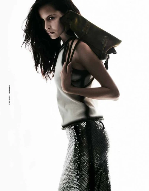 Fashion editorial shot of a woman in a white top and sequined skirt with a leather bag slung over her shoulder against a white backdrop.