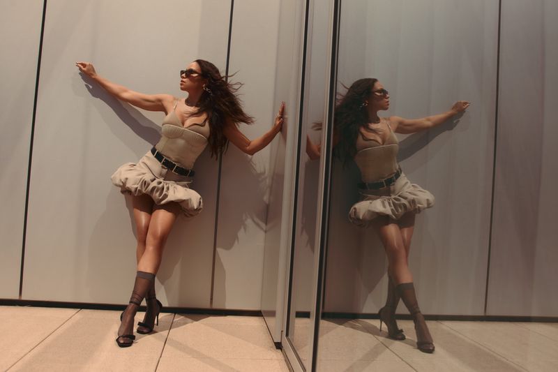 A woman in a tan dress poses fashionably with arm extended by a reflective panel, reflecting her image; she wears heels and dark sunglasses.