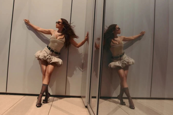 A woman in a tan dress poses fashionably with arm extended by a reflective panel, reflecting her image; she wears heels and dark sunglasses.