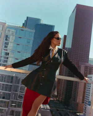 Jurnee Smollett leaning on a balcony railing against a backdrop of urban skyscrapers wearing a dark jacket and red skirt