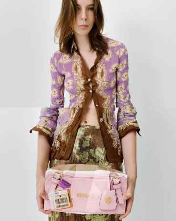 A woman models a floral-patterned jacket and floral skirt while holding a pink Versace handbag in a studio with a white background.