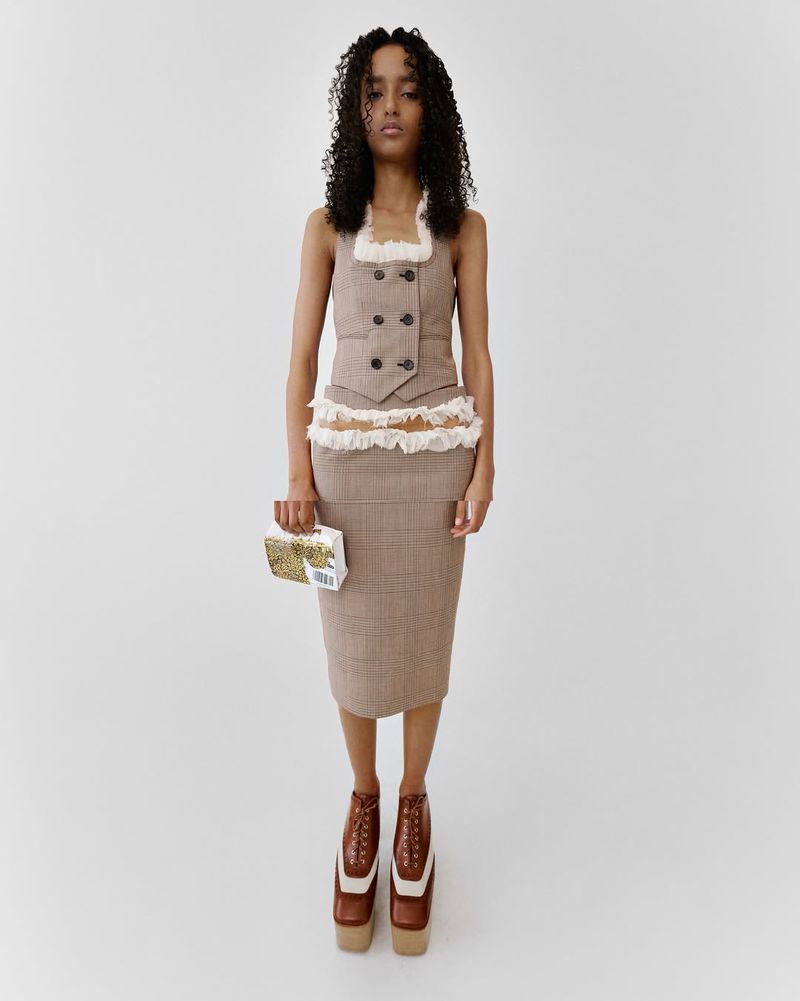 A tan-skinned woman with curly hair poses in a double-breasted vest, plaid skirt, platform heels, and holds a patterned clutch against a solid white background.