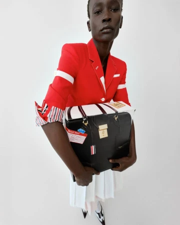 Woman in red blazer holds a black and white travel bag, posing in a fashion shot on a white background. Pleated skirt and heels visible.