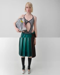 Model with blonde hair wearing a grey cutout top and green pleated skirt holding a silver parcel against a grey background.
