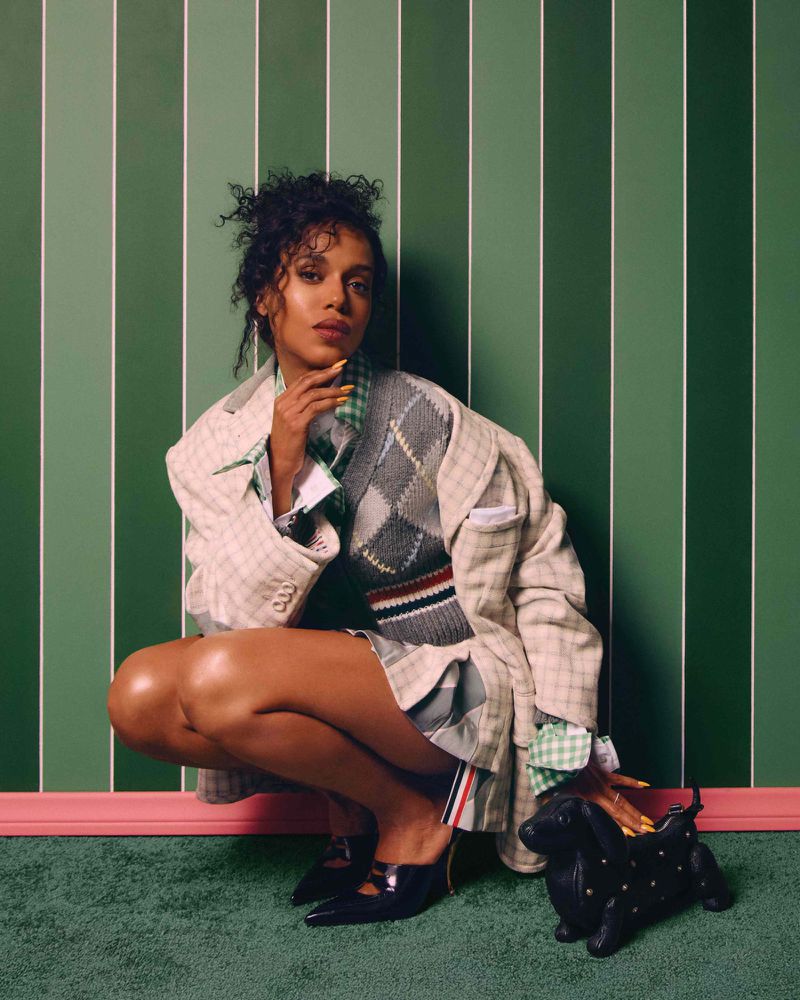 A young woman crouches indoors next to a Dachshund-shaped purse, wearing a plaid blazer, argyle vest, and black heeled shoes in a studio setting.