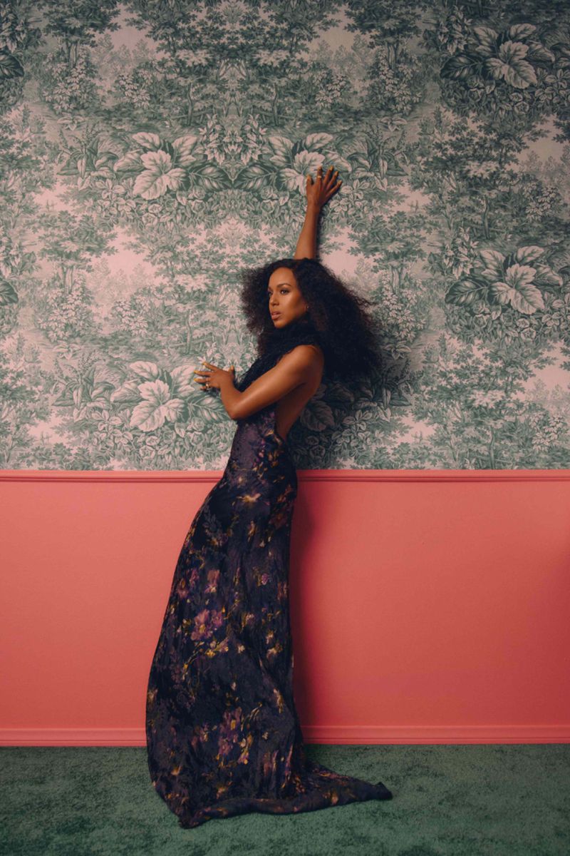 Elegant woman poses in floral gown against a patterned wallpaper backdrop, arms extended, capturing a blend of fashion and artistic design.