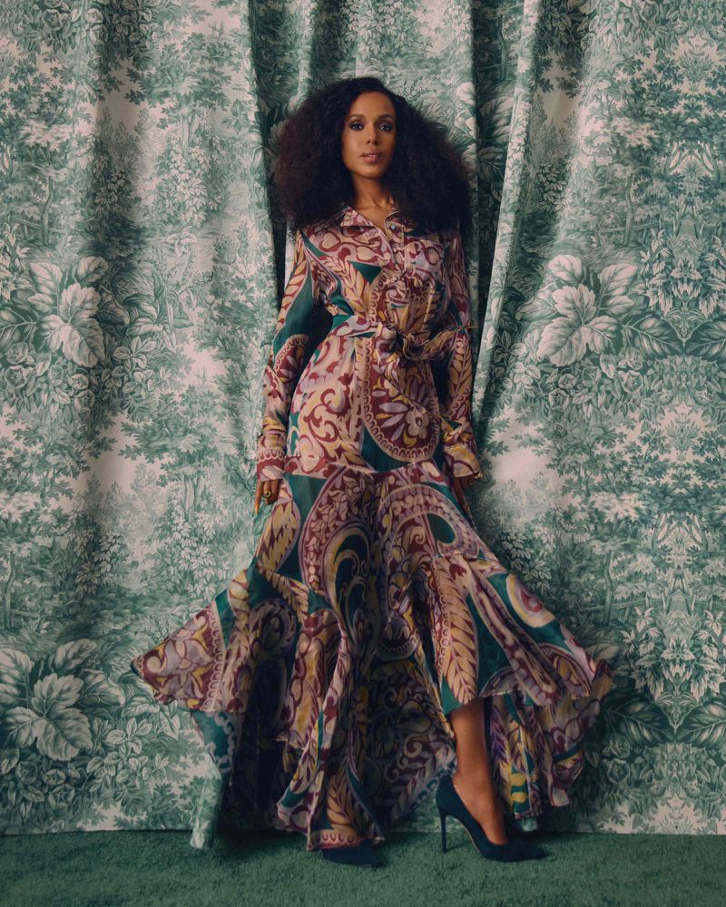 Elegant adult woman poses in a paisley print dress and high heels against a green toile patterned curtain backdrop in a studio setting.