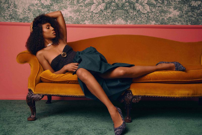 Adult woman with curly hair in an off-the-shoulder dress and heels, reclining on a yellow velvet couch in an indoor setting.