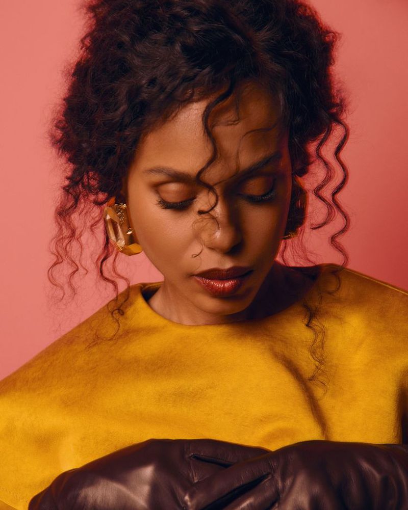 Portrait of a curly-haired woman with dark gloves and a yellow top against a reddish-pink backdrop in a studio setting.