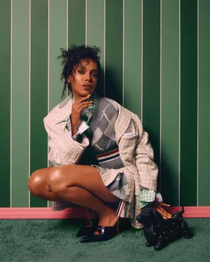 Kerry Washington squatting against a vertical striped green wall wearing a plaid blazer, sweater vest, and resting a hand on a dog-shaped handbag