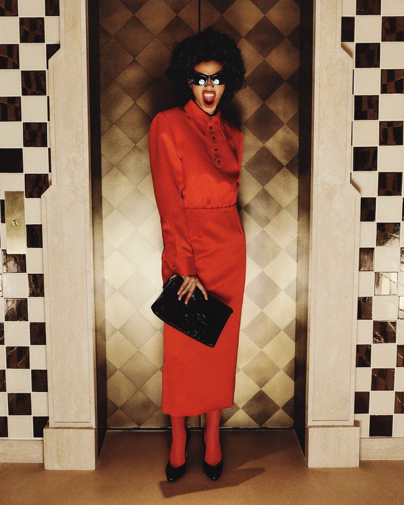 A woman with an afro and sunglasses stands in front of an elevator in a red skirt suit, holding a black clutch purse.