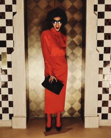A woman with an afro and sunglasses stands in front of an elevator in a red skirt suit, holding a black clutch purse.