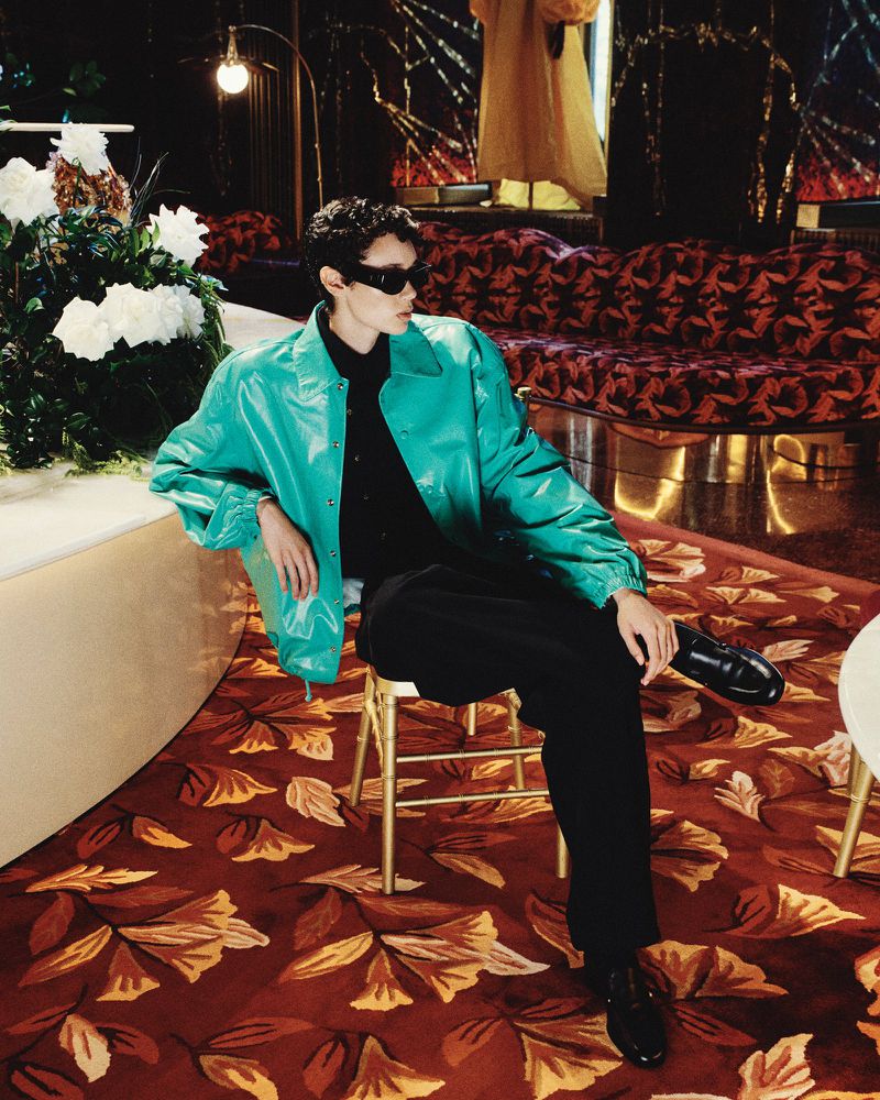 Stylish adult in sunglasses and a blue jacket sits in an interior lounge with a leaf-patterned carpet and decorative floral arrangements.