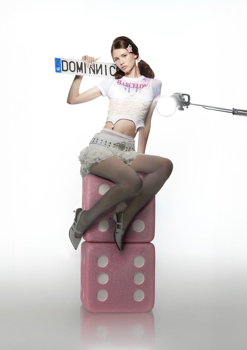 Young adult female model sitting on stacked pink dice in a photo studio, holding a license plate. She wears white top and skirt.