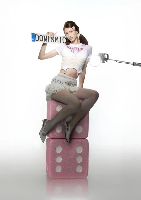 Young adult female model sitting on stacked pink dice in a photo studio, holding a license plate. She wears white top and skirt.