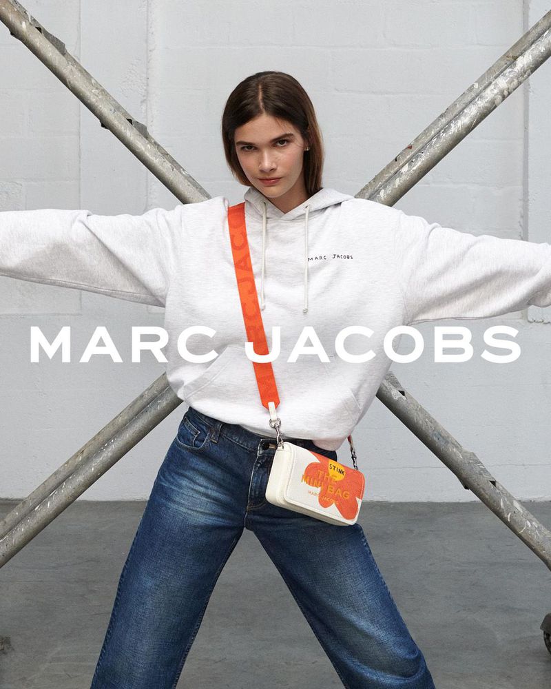 A brunette woman poses indoors wearing a gray hoodie, blue jeans, and a small Marc Jacobs flower-themed purse on a light gray background.