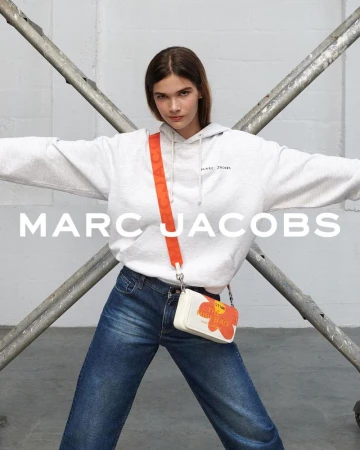A brunette woman poses indoors wearing a gray hoodie, blue jeans, and a small Marc Jacobs flower-themed purse on a light gray background.