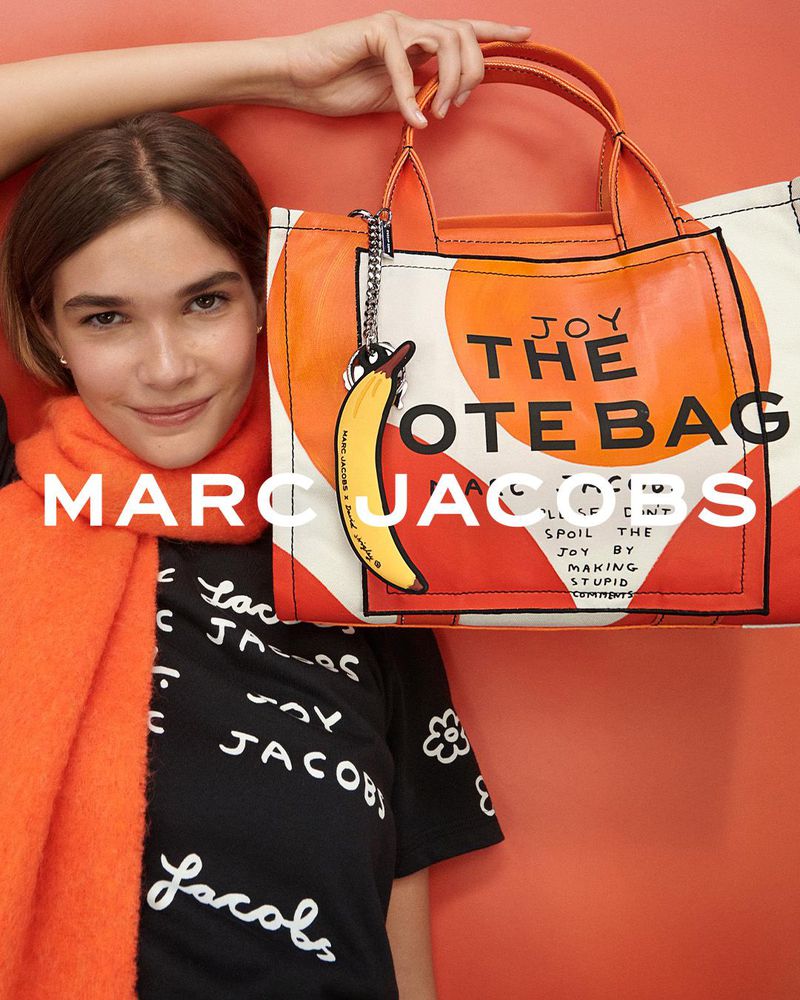 Woman poses with a Marc Jacobs tote bag and scarf against an orange background, showcasing the brand's latest fashion accessory.