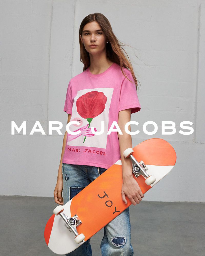 A woman poses indoors against a white wall, showcasing a pink graphic tee, patchwork jeans, and holding an orange and white skateboard.