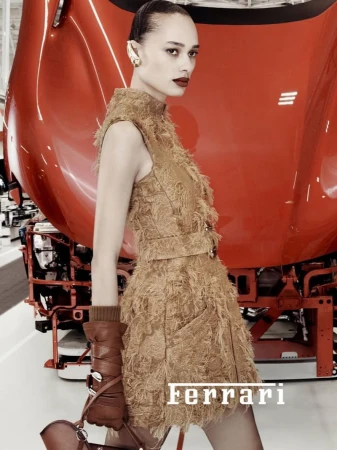 Fashion shot of a woman posing in front of a bright red car, wearing a tan dress, brown gloves and carrying a brown handbag.