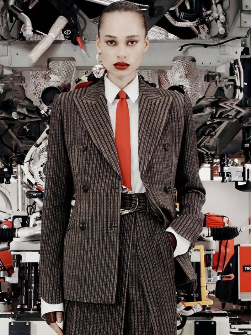 Adult woman in a pinstripe suit, white shirt, and red tie poses in front of industrial equipment for a fashion editorial shoot.