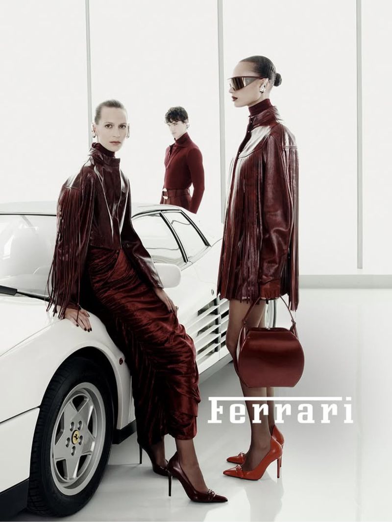 Fashion editorial shot of three models in burgundy outfits with a white Ferrari. One model is leaning on the car, one is standing, and one is behind the car.