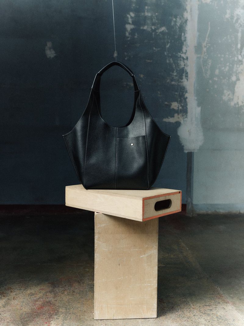 Black leather handbag on a wooden pedestal against a textured gray and white background, studio lighting.