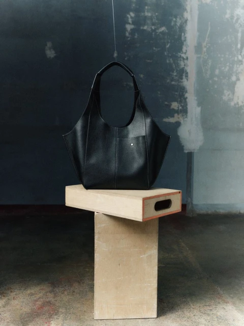 Black leather handbag on a wooden pedestal against a textured gray and white background, studio lighting.