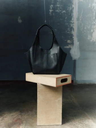 Black leather handbag on a wooden pedestal against a textured gray and white background, studio lighting.