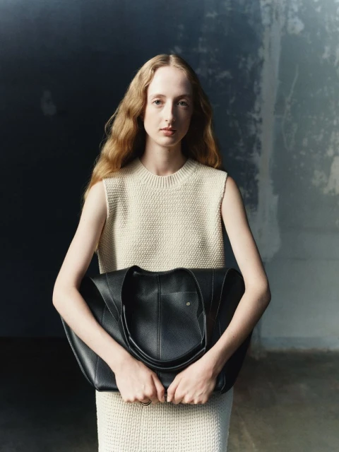 Woman in cream dress holding black tote bag in front of a mottled grey wall with light on the model's face and wavy hair.