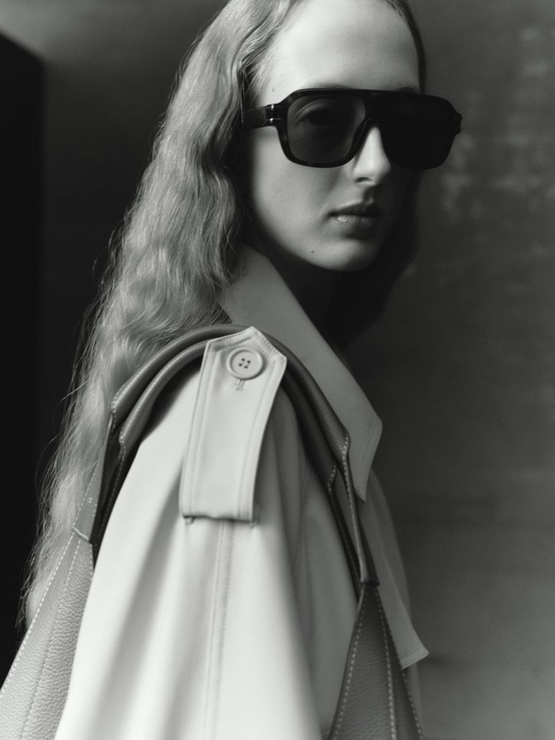 Monochrome studio shot of a blonde adult woman wearing dark sunglasses and a light-colored coat, posing with a serious expression.