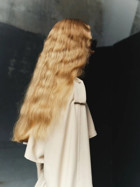 Woman with long blonde wavy hair and sunglasses, wearing a beige trench coat, stands in an indoor setting against a dark background.